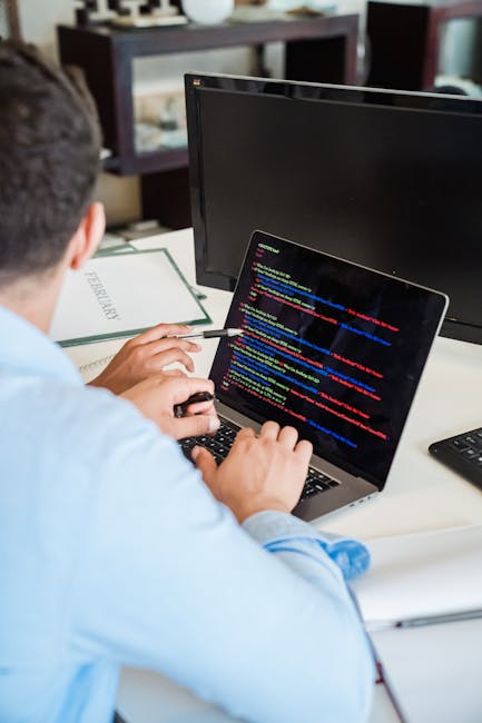 A person seated at a desk, viewed from behind, working on a laptop with a dark screen displaying colorful lines of computer code. The individual, wearing a light blue shirt, has short dark hair and is