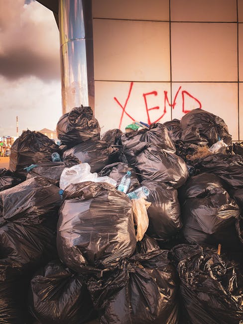 A large pile of black bin bags filled with rubbish is stacked against a tiled wall in an outdoor urban setting. Some of the bags are partially torn, revealing discarded plastic bottles and other waste